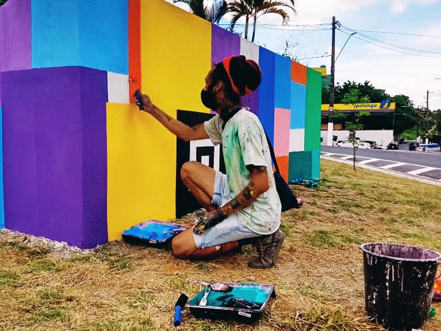 imagem mostra o artista Kadu colorindo utilizando as cores roxo, azul, rosa, verde, laranja, amarelo, preto e branco o muro do canteiro na avenida souza ramos, 